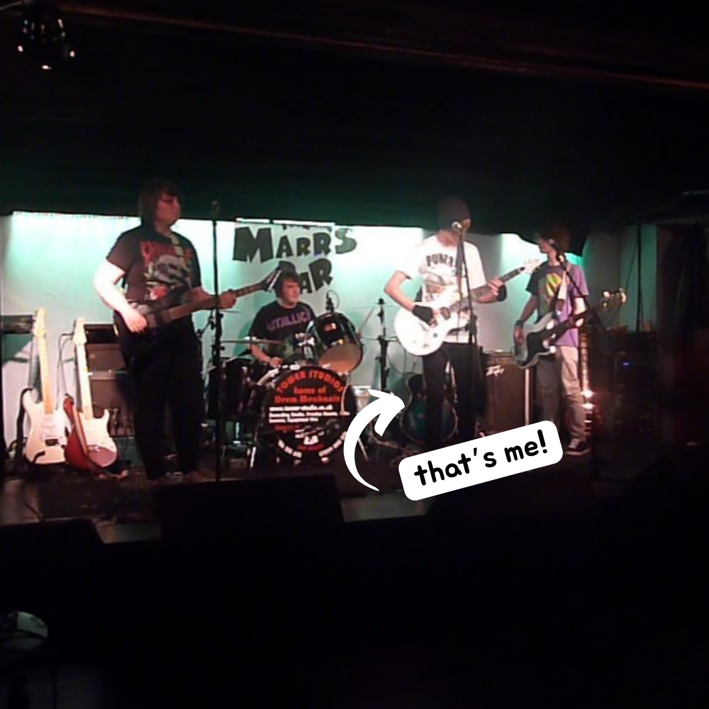 A grainy photograph of Luke on stage at 'The Marrs Bar' playing guitar in his band. He is notably wearing a Funeral for a Friend t shirt, black beanie hat, black fingerless gloves and has a straightened fringe.