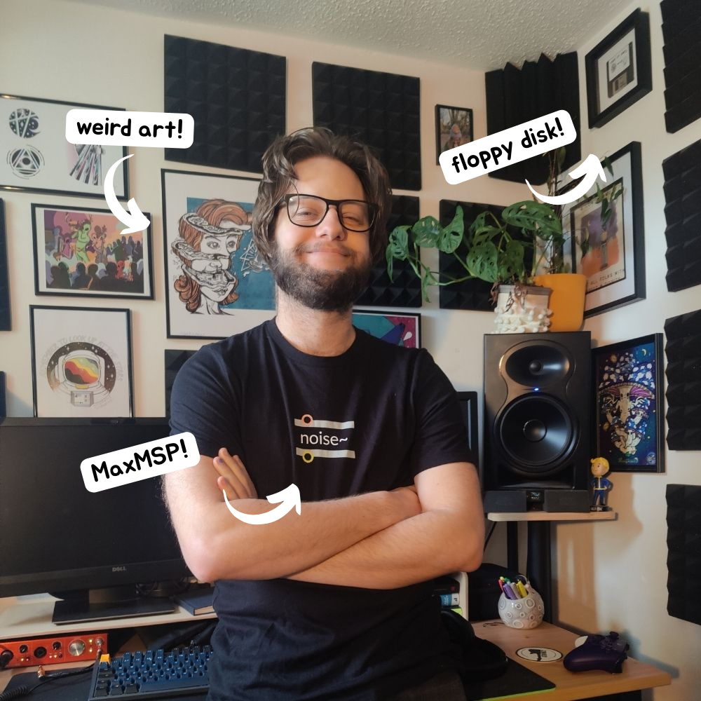 A smiley Luke stands proudly next to his desk with his arms crossed in a power pose. He is wearing a Max MSP t shirt with the 'noise~' object on it.