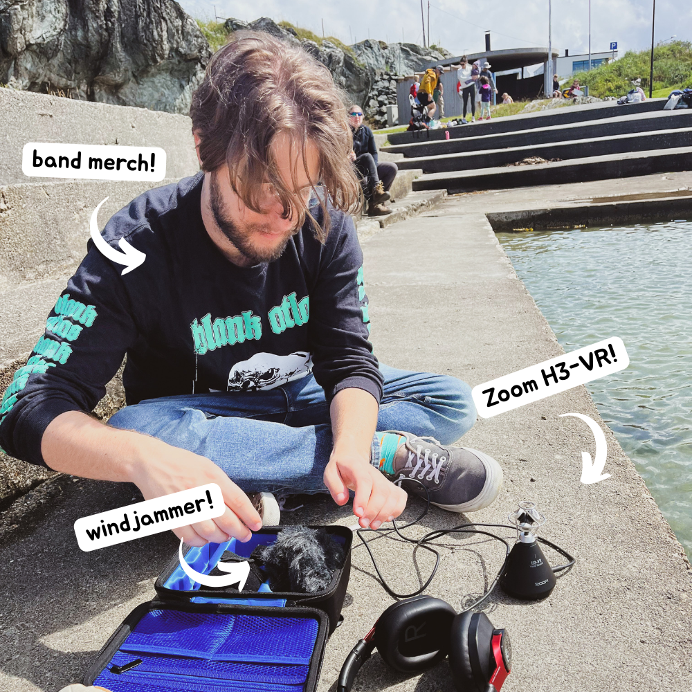 Luke sat next to a body of water setting up a Zoom H3-VR microphone. Text popouts point to different picture features. #1 points to Luke's t-shirt for the band Blank Atlas and reads 'band merch!'. #2 points to a microphone wind coer and reads 'windjammer!'. #3 points to Luke's recorder and reads 'Zoom H3-VR!'
