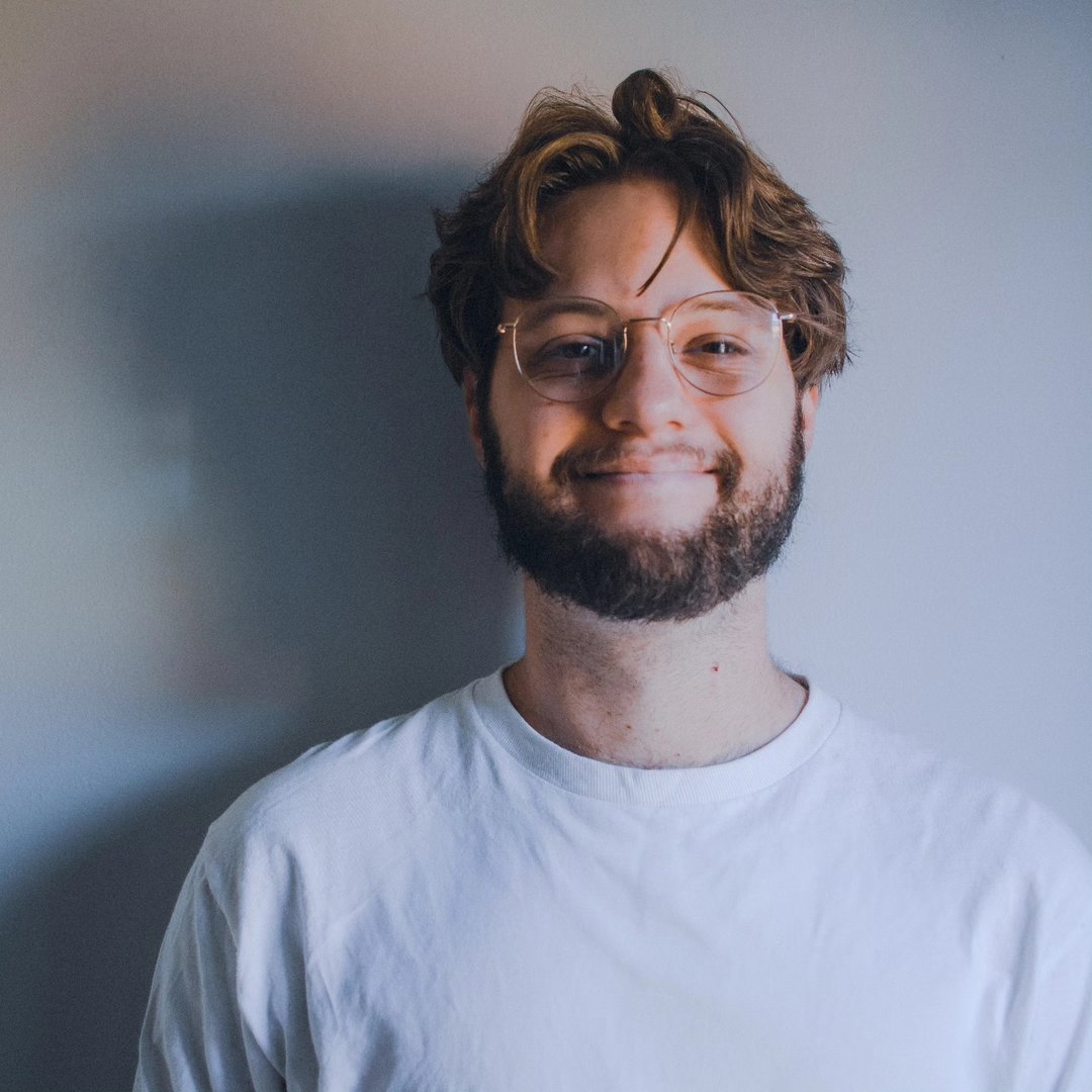 A portrait of Luke. He has medium-length, scruffy, middle-parted hair, a short beard, rounded gold-frame glasses and big ol' smile.