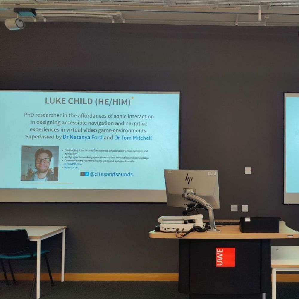A Powerpoint presentation projected on a screen in a classroom. The key text content reads: 'Luke Child (He/Him) - PhD Researcher in the affordances of sonic interaction in designing accessible navigation and narrative experiences in virtual video game environments. Supervised by Dr Natanya Ford and Dr Tom Mitchell.